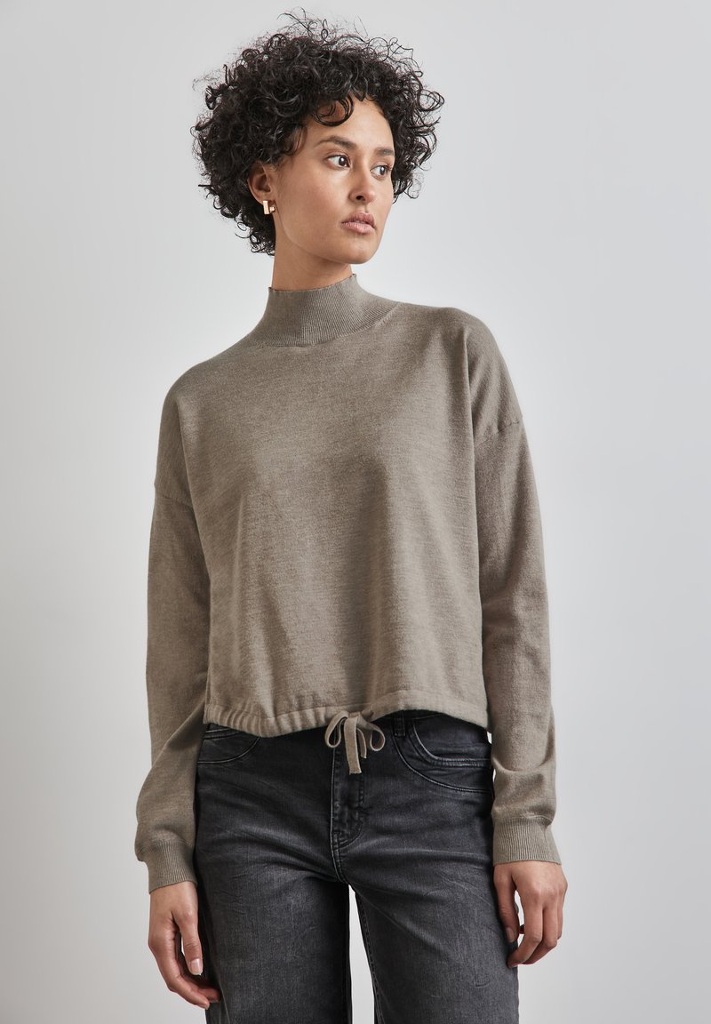 Maglione corto di colore marrone chiaro con collo alto e orlo con cordoncino. Texture morbida, maniche lunghe e polsini a coste si abbinano bene a jeans neri casual.