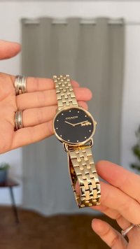 Gullfarget klokke med rund svart skive, sølvfargede time markører, og lenkebånd. Merkevaren er tydelig vist på skiven.