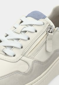 Αθλητικό παπούτσι από κρεμ δέρμα με ανοιχτό γκρι λεπτομέρειες, διαθέτει λευκά κορδόνια, μια τσέπη στο πλάι με φερμουάρ και μια μπλε υφασμάτινη λεπτομέρεια στη φτέρνα.