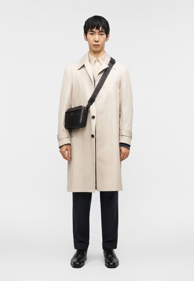 Μπεζ παλτό με δομημένο σχεδιασμό, φορεμένο πάνω από ένα ανοιχτόχρωμο πουκάμισο και παντελόνια με λεπτές ρίγες. Μαύρη τσάντα χιαστί με λογότυπο, γυαλιστερά παπούτσια.