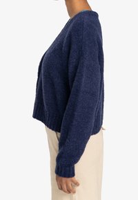 Marinblå stickad tröja, kort design, långa ärmar, texturerat tyg, ribbad fåll, bärs med beige byxor.