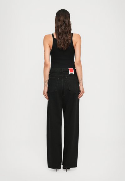 Woman with long wavy hair wears a black tank top and high-waisted wide-leg black jeans with a red and white label on the waistband.