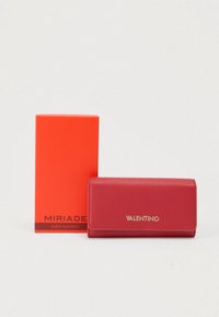 Κόκκινο πορτοφόλι από δερμάτινη επιφάνεια με υφή, με χρυσό λογότυπο, δίπλα σε πορτοκαλί κουτί. Το πορτοφόλι διαθέτει κλείσιμο με καπάκι και ορθογώνιο σχήμα.