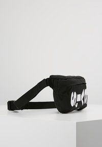 Svart tygmidjeväska med justerbar rem och vit fet deltext framtill, placerad på en vit yta mot en enkel bakgrund.