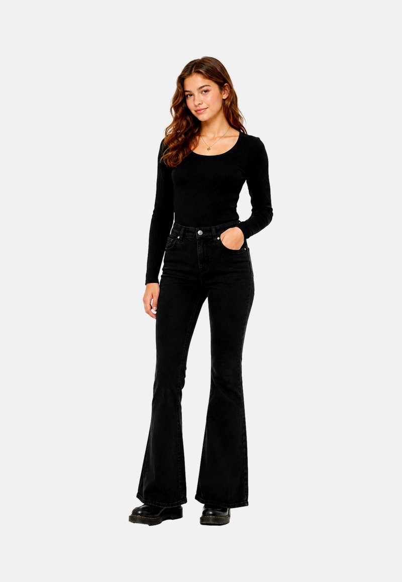 Jeune femme aux longs cheveux bruns portant un haut noir à manches longues, un jean noir taille haute évasé et des bottes noires, debout avec une main dans la poche.