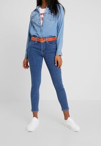 Camisa de mezclilla azul celeste con diseño de botones, usada sobre una camiseta blanca con gráfico, combinada con jeans ajustados azul oscuro y zapatillas blancas.