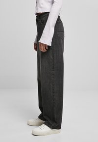 Jeans noirs à jambe large en denim, avec une taille haute et un design classique à cinq poches, associés à des baskets blanches.