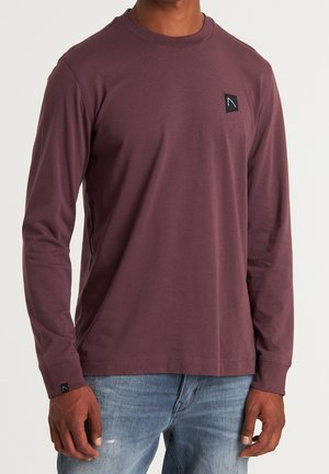Homme portant une chemise bordeaux à manches longues avec des écussons noirs sur la poitrine et le poignet, associée à un jean bleu clair.
