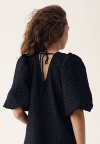 Femeie cu păr castaniu ondulat purtând o rochie neagră texturată cu mâneci bufante și spate decupat legat cu bretele subțiri.