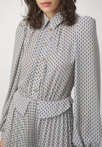 Blusa a maniche lunghe bianca e nera con motivo, colletto, chiusura con bottoni e design plissettato con bottoni dorati e vita legata.