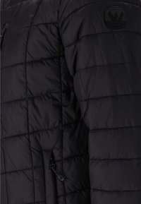 Sort pufferjakke med quiltet tekstur, med en lynlåslomme og et rundt logo på skulderen. Materialet ser skinnende og let ud.