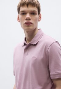 Svetlo roza polo majica s tremi gumbi, rebrastim ovratnikom in subtilno vezenim logotipom. Gladek material s kratkimi rokavi in teksturiranimi poudarki.