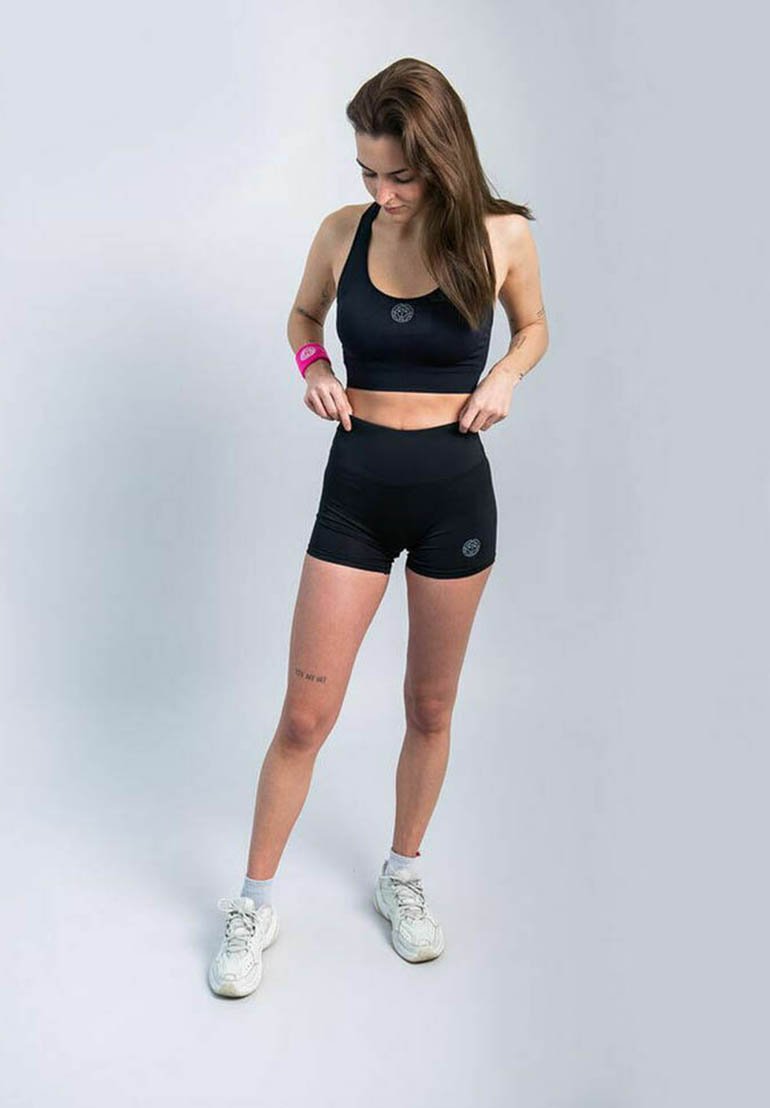 Top crop atletico nero con logo e pantaloni corti a vita alta coordinati, abbinati a sneakers bianche e un braccialetto rosa. Tessuto liscio e elasticizzato.