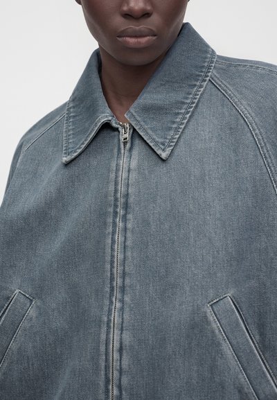 Gros plan d’une personne portant une veste en jean bleue délavée avec un large col, une fermeture éclair à l’avant et des poches latérales inclinées, sur un fond uni.