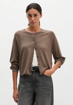 Frau mit dunklem Haar trägt eine braune, geknöpfte Strickjacke über einem weißen Top und dunkelgraue Jeans mit schwarzem Gürtel, steht mit einer Hand in der Tasche.