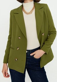 Oliwkowo-zielona dwurzędowa marynarka z wyciętymi klapami, dwoma przednimi kieszeniami i okrągłymi guzikami. Nosi się ją na kremowy golf i ciemne dżinsy.