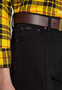 Persona che indossa jeans neri con etichetta Polo, una cintura in pelle marrone e una camicia a quadri gialla con strisce nere e rosse.