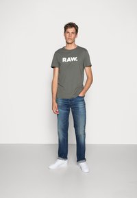 Zelena majica s bijelim logotipom "RAW.", uparena s plavim trapericama i bijelim tenisicama. Jednostavan dizajn, kratki rukavi, opušteni kroj.