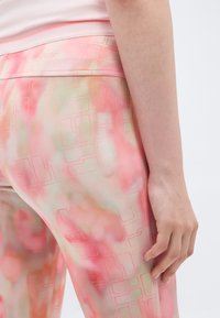 Zbliżenie ręki osoby spoczywającej obok biodra, ubranej w różowe spodnie w jasny wzór tie-dye z detalami geometrycznymi.