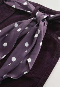 Tissu en velours côtelé violet avec une écharpe lisse et à motifs, ornée de pois blancs, nouée autour de la taille. Aucun détail supplémentaire présent.