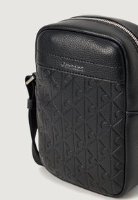 Melna ādas pārsējošā soma ar teksturētu virsmu, kurai ir ģeometrisks raksts un sudraba logo. Augšējā daļā slēdzama ar rāvējslēdzēju.