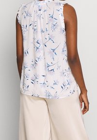Blusa floreale in tessuto leggero con motivi di fiori blu e rosa, maniche a rouches e scollatura arricciata. Abbinata a pantaloni beige.