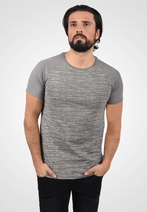 Camiseta gris de manga corta con textura jaspeada, con cuello redondo y mangas raglán en contraste. Llevada con las manos en los bolsillos.