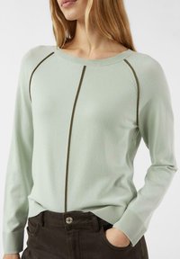 Kobieta ubrana w jasnooliwrowy sweter z ciemnymi pionowymi i barkowymi paskami, w ciemnych spodniach, z ręką w kieszeni, na białym tle.