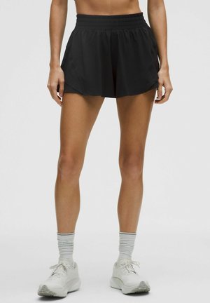 Frau trägt schwarze Sportshorts, weiße Socken und weiße Laufschuhe, steht vor einem einfarbigen Hintergrund.