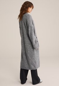 Garšs pelēks kardigāns mīkstā, teksturētā adījumā. Piedāvā kritušas plecus, ribota apkakle un ceļu garuma dizainu. Saskaņots ar tumšiem biksēm.