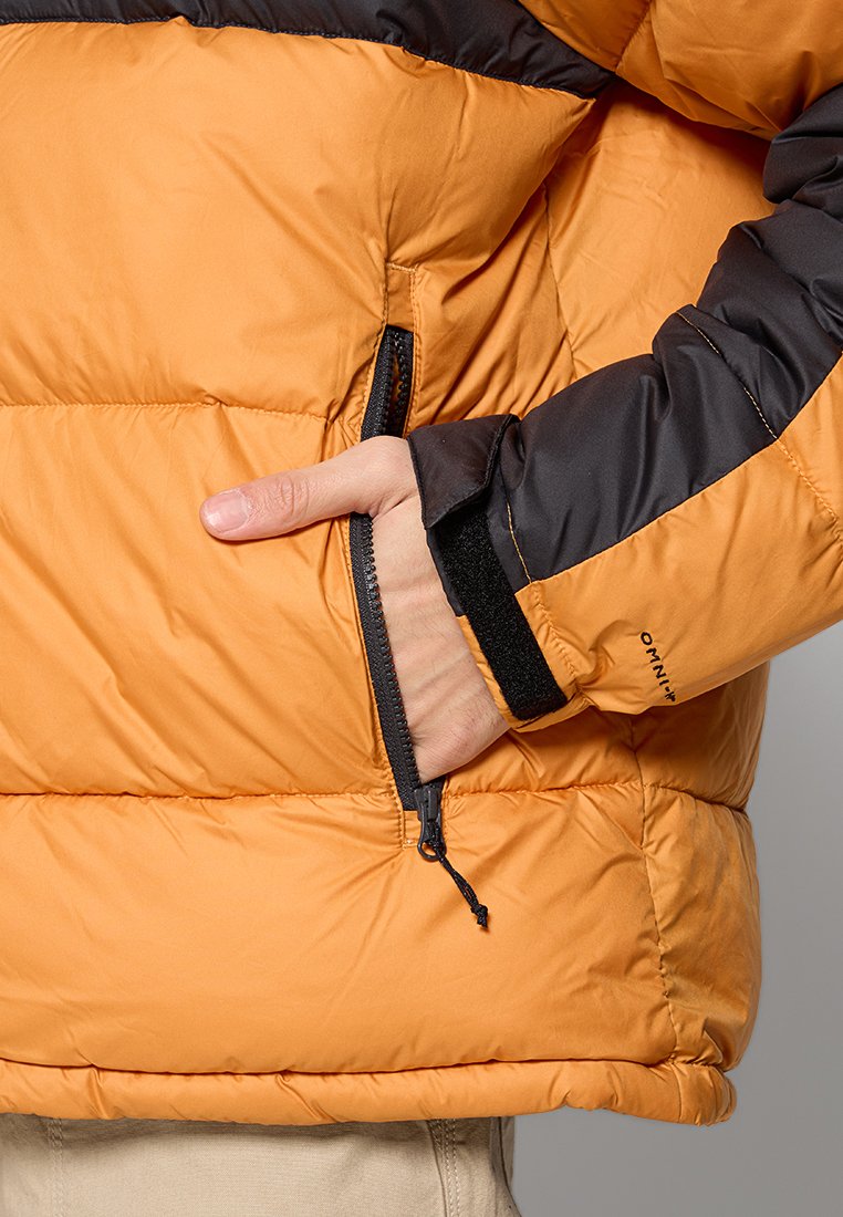 Daunenjacke mit orangefarbenem Körper und schwarzen Ärmeln, ausgestattet mit einer Reißverschlusstasche und einem Klettverschluss an den Ärmelabschlüssen. Strukturierter Stoff mit gestepptem Design.