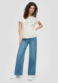 Kurze Ärmel cremefarbenes T-Shirt mit kreisförmigem grünem Grafiktext, kombiniert mit weit geschnittenen blauen Denim-Jeans und weißen Sneakers.