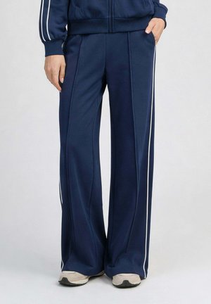 Personne portant un pantalon de jogging bleu marine à jambes larges avec des bandes blanches sur les côtés et des baskets blanches, debout les mains dans les poches.