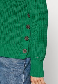 Pull en tricot vert avec un design texturé, mettant en avant de grands boutons foncés sur le côté et un petit accent logo sur la manche.