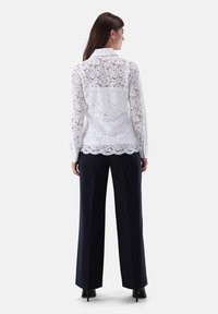 White lace blouse with floral patterns, collared design, long sleeves, and scalloped hem, paired with dark wide-leg trousers.