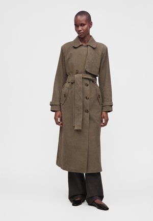Femme portant un long trench-coat vert olive ceinturé avec de gros boutons et un large col, debout avec une posture droite sur un fond uni.