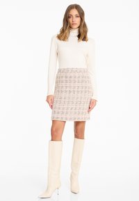 Brede gebreide coltrui in crème gecombineerd met een getextureerde kokerrok met een meerkleurig ruitpatroon; kniehoge beige laarzen.