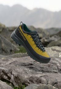 La Sportiva vaelluskengät tumman vihreänä ja keltaisena, joissa on teksturoitu päällinen, nauhat ja kestävä kumipohja, joka on suunniteltu ulkokäyttöön.