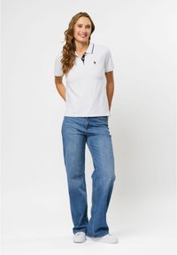 Witte polo met korte mouwen en een marineblauwe kraag en logo, gecombineerd met lichtblauwe flared jeans en witte sneakers, staand tegen een effen achtergrond.