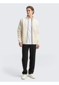 Witte textuur ritsjack met hoge kraag, gedragen over een eenvoudige witte T-shirt en gecombineerd met zwarte broek en witte sneakers.