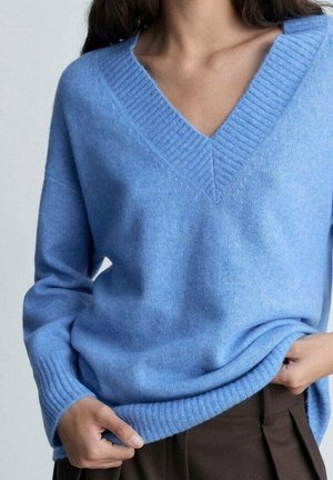 Femme portant un pull bleu clair à col en V avec col et poignets côtelés, tenant l'ourlet, associé à un pantalon marron foncé.