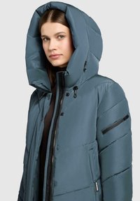 Kvinde iført en teal vinterjakke med polstring, der har en overdimensioneret hætte og en sort lynlåslomme på den øverste ærme.