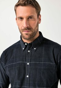 Hombre con cabello corto castaño y barba, vistiendo una camisa negra de botones con un sutil patrón de cuadros, mirando al frente contra un fondo liso.