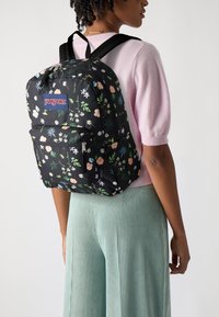 Juoda audinio kuprinė su pastelinių spalvų gėlių raštu, turinti priekinį kišenę ir reguliuojamus diržus, pažymėta "Jansport" logotipu.