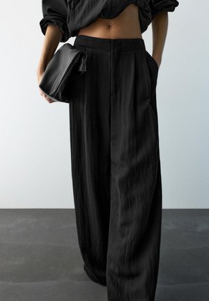 Osoba ubrana w luźne, czarne, wysokie spodnie i krótki czarny top, trzymająca złożoną czarną kopertówkę na jednolitym tle.