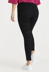 Person som bär tighta svarta jeans, en rosa kortärmad topp och vita sneakers, som går bortåt mot en helt vit bakgrund.