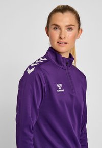 Lila zip-jacka i slätt tyg med vitt logotyp och chevronaccenter på axlarna. Hög krage och figurnära design.