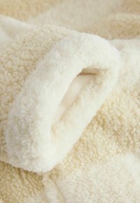 Krämfärgad filt med en mjuk, fluffig textur. Har en slät kant, där den ena sidan har ett kort, plyschigt tyg och den andra en mysig yta.