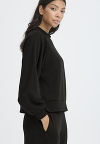 Melnā sporta krekls ar apaļu izgriezumu un garām, apjomīgām piedurknēm. Iespējas ietver ribotas piedurknes un brīvu piegriezumu. Mīksta, gluda tekstūra.