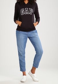 Sweat à capuche noir avec imprimé « GAP », poches frontales, assorti à un jean bleu clair et des chaussures blanches à enfiler. Tenue décontractée avec une coupe confortable.